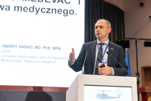 Elegancko ubrany mężczyzna w czasie wystąpienia. Stoi za mównicą. Prawą rękę ma uniesioną.