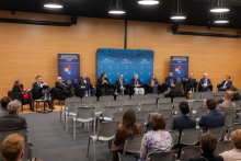 Panel dyskusyjny w sali konferencyjnej, w którym uczestniczy dwanaście osób siedzących w czarnych fotelach ustawionych wzdłuż drewnianej ściany. Przed każdym fotelem znajdują się małe stoliki z butelkami wody i szklankami. W tle widoczna jest niebieska ścianka z napisami informującymi o jubileuszach.