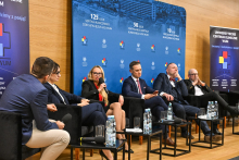 Sześć osób siedzących w czarnych fotelach ustawionych w rzędzie na scenie w sali konferencyjnej. Przed każdym fotelem znajdują się małe stoliki z butelkami wody i szklankami. Jedna z osób trzyma mikrofon, co wskazuje na trwającą dyskusję