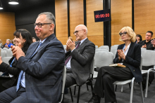 Kilka osób siedzących w rzędach na krzesłach w sali konferencyjnej, zwróconych w stronę sceny. Osoby mają na sobie eleganckie stroje biznesowe i trzymają dłonie w geście oklasków. 