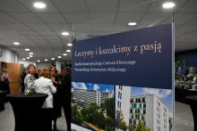 Plakat z napisem „Leczymy i kształcimy z pasją. Benefis Uniwersyteckiego Centrum Klinicznego Warszawskiego Uniwersytetu Medycznego” oraz dwoma zdjęciami nowoczesnych budynków. W tle osoby stojące przy wysokich stołach podczas wydarzenia w jasnej sali z oświetleniem sufitowym.