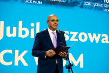 Osoba w garniturze z krawatem stoi przed niebieskim tłem z białym napisem, trzyma teczkę i mówi do mikrofonu, prawdopodobnie podczas uroczystości jubileuszowej.