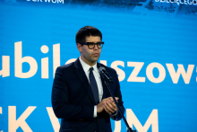 Mężczyzna w okularach, ciemnym garniturze, białej koszuli i krawacie stoi przed mikrofonem, trzyma dłonie złożone. W tle niebieskie tło z białym napisem 