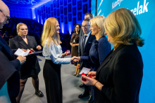 Kilka osób w stroju formalnym stoi na scenie, wymieniając uściski dłoni i odbierając małe czerwone pudełka, prawdopodobnie z odznaczeniami.