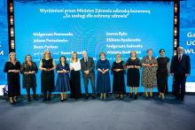 Grupa osób w stroju formalnym stoi na scenie przed niebieskim tłem z napisem „Wyróżnieni przez Ministra Zdrowia odznaką honorową ‘Za zasługi dla ochrony zdrowia’” oraz listą nazwisk. Uczestnicy trzymają małe czerwone pudełka, prawdopodobnie z odznaczeniami.