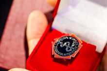 Zbliżenie na dłoń trzymającą otwarte czerwone pudełko z odznaką w kształcie gwiazdy, z czarnym okrągłym środkiem i srebrnym napisem.