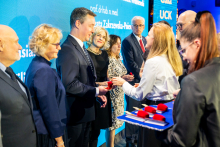 Grupa osób w strojach formalnych na scenie podczas gali UCK. Na pierwszym planie kobieta w białej koszuli wręcza czerwone etui mężczyźnie w garniturze