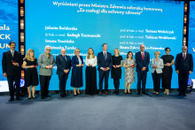 Grupa osób w stroju formalnym stoi na scenie przed niebieskim tłem z napisem „Wyróżnieni przez Ministra Zdrowia odznaką honorową ‘Za zasługi dla ochrony zdrowia’” oraz listą nazwisk. Uczestnicy trzymają małe czerwone pudełka, prawdopodobnie z odznaczeniami.