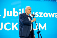 Osoba w ciemnym garniturze z muszką stoi przy mikrofonie i przemawia. W tle jasnoniebieski ekran z dużym białym napisem