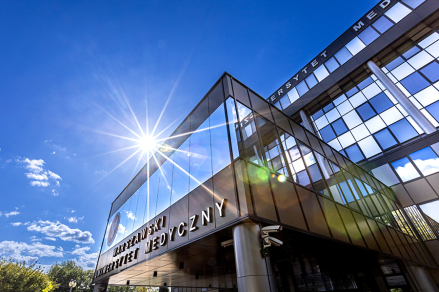 Nowoczesny budynek Warszawskiego Uniwersytetu Medycznego w słoneczny dzień. Na pierwszym planie szklana fasada z napisem „Warszawski Uniwersytet Medyczny”. W tle niebieskie niebo z białymi chmurami i rozbłysk słońca na szkle. Po lewej stronie widoczne są drzewa i krzewy.