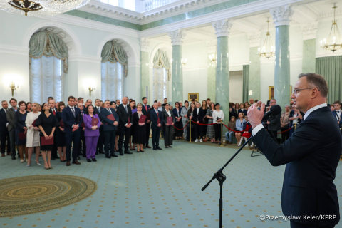 Grupa osób stojąca w dużej sali znajdującej się w Pałacu Prezydenckim. Wszystkie osoby są elegancko ubrane. Po prawej elegancko ubrany mężczyzna - Prezydent RP. Prezydent przemawia do mikrofonu. Ma uniesioną rękę. 