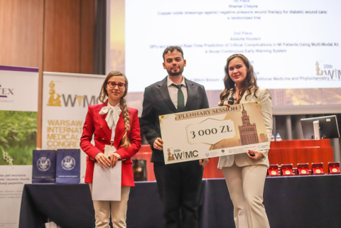 Trzy osoby. Stoją na środku auli podczas ceremonii wręczenia nagród. Po lewej i po prawej młode kobiety, w środku - młody mężczyzna. Kobieta po lewej ma na sobie czerwoną marynarkę i trzyma dokumenty. Mężczyzna w środku ma na sobie ciemny garnitur i krawat. Kobieta po prawej ma na sobie jasną marynarkę i trzyma duży czek o wartości 3000 złotych z napisem "20th WIMC". 