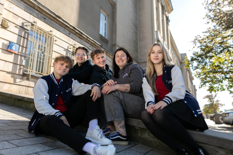 Pięć młodych osób: dwie kobiety i trzech mężczyzn. Uśmiechają się i patrzą przed siebie. Siedzą na schodach przed szarym budynkiem. Budynek ma duże okna. Dwie osoby po lewej stronie noszą granatowe bluzy z białymi rękawami i napisem WUM. Osoba po prawej stronie również ma taką kurtkę. Pozostałe dwie osoby są ubrane w ciemne ubrania. W tle widać drzewa i niebo.