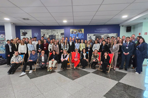 Grupa około czterdziestu osób pozujących do wspólnego zdjęcia. Część osób stoi, a część siedzi na krzesłach w dwóch rzędach. Wszyscy uśmiechają się do zdjęcia. Część osób jest ubrana formalnie, część nieformalnie. W tle znajdują się plakaty związane z medycyną. Pomieszczenie jest jasno oświetlone. Sufit jest podwieszany, a podłoga wyłożona szarymi płytkami.