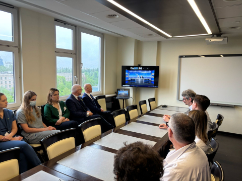 Grupa osób siedzi przy długim stole w sali konferencyjnej, oglądając prezentację wyświetlaną na ekranie telewizora. W pomieszczeniu znajduje się tablica suchościeralna, a za oknami widoczny jest zielony krajobraz i budynki.