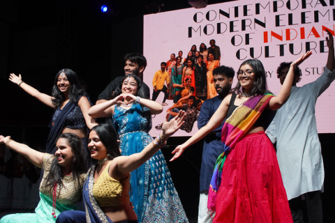 Grupa młodych osób - studentów WUM - stojących na scenie. Mają na sobie kolorowe, tradycyjne stroje. Wykonują różne gesty rękami i pozują do zdjęcia. W tle znajduje się duży ekran z napisem „Contemporary modern elements of Indian culture”