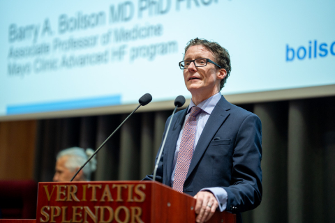 Mężczyzna w średnim wieku, w okularach. Ma na sobie ciemny garnitur, jasnoniebieską koszulę i jasnoróżowy krawat. Stoi za mównicą i przemawia. Mównica jest brązowa i ma napis "VERITATIS SPLENDOR" w kolorze złotym. W tle znajduje się ekran multimedialny z tekstem.