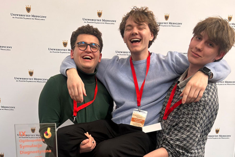 Trzech młodych mężczyzn - studentów WUM na tle ścianki z napisem „Uniwersytet Medyczny im. Piastów Śląskich we Wrocławiu”. Mężczyzna w środku jest trzymany na rękach przez mężczyzn stojących po prawej i po lewej. Wszyscy się uśmiechają. Na szyjach maja czerwone smycze z identyfikatorami. Osoba po lewej stronie ubrana jest w zielony sweter, osoba w środku w niebieski sweter, a osoba po prawej stronie ma na sobie szarawy sweter. Na pierwszym planie znajduje się nagroda z napisem „Symulacje Diagnostyczne”.