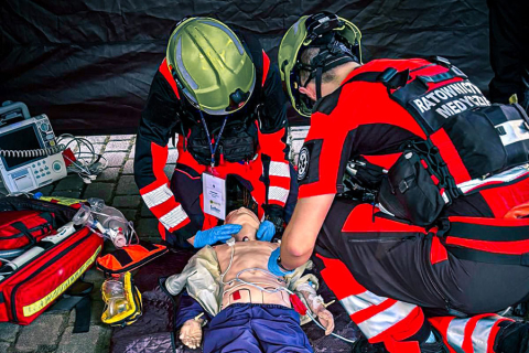 Dwóch ratowników medycznych w czerwono-czarnych uniformach i zielonych kaskach wykonuje resuscytację na manekinie. Jeden prowadzi masaż serca, drugi udrażnia drogi oddechowe. Obok leży sprzęt medyczny. Scena odbywa się na zewnątrz, pod czarnym namiotem.