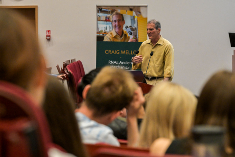Prof. Craig C. Mello, mężczyzna w żółtej koszuli, z mikrofonem stoi przed publicznością w sali wykładowej. Gestykuluje ręką. W tle plakat z napisem ‘CRAIG MELLO RNA: Ancient Mechanisms’.