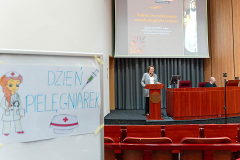 The interior of the auditorium with wooden panels on the walls and rows of red armchairs. In the foreground, to the left, a poster with the caption “NURSES' DAY” is visible. The poster features a drawing of a nurse in a white uniform, with a stethoscope around her neck and a nurse's cap with a red cross. Next to the caption is an illustration of a syringe. In the depth of the photo, on the stage, stands a woman speaking from behind a lectern. She is wearing a light-colored jacket, glasses and beads.