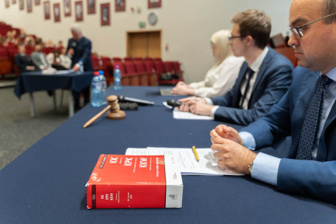 Sala. Kilka osób siedzi przy stole przykrytym granatowym obrusem. Na stole znajdują się różne przedmioty, w tym duża czerwona książka z białym napisem 'KK KPK KKW', młotek sędziowski, butelki wody, długopis oraz dokumenty. W tle widać publiczność siedzącą na czerwonych krzesłach oraz mężczyznę stojącego przy innym stole i mówiącego do mikrofonu.