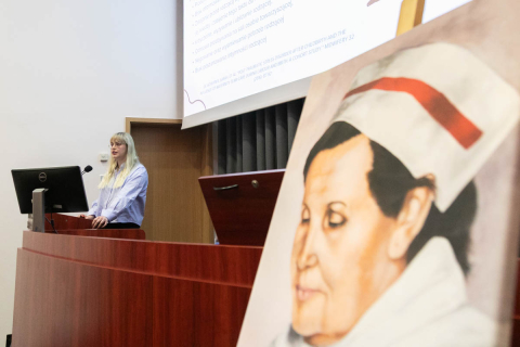 Kobieta stojąca za drewnianą mównicą w sali wykładowej. Ubrana jest w jasnoniebieską koszulę, ma jasne włosy. Stoi przed komputerem oraz mikrofonem. W tle widoczny jest ekran projekcyjny z częściowo czytelnym tekstem. Na pierwszym planie, po prawej stronie, znajduje się plakat przedstawiający twarz kobiety z białym czepkiem, na którym jest czerwony pasek.