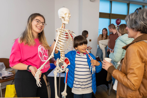 Grupa osób w pomieszczeniu. Na pierwszym planie  kobieta w różowej koszulce z białym logo, stojąca obok dziecka w niebieskiej kurtce i koszulce w paski. Oboje trzymają i oglądają model ludzkiego szkieletu.