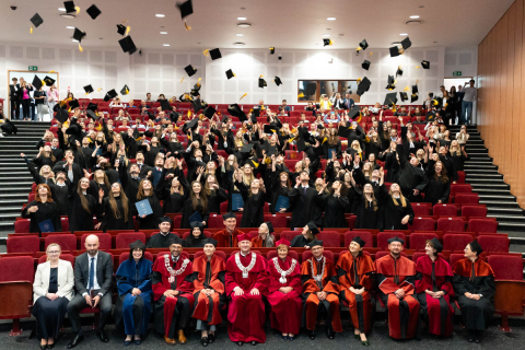 Uroczystość rozdania dyplomów w dużej, jasno oświetlonej auli. W centrum znajduje się grupa absolwentów ubranych w tradycyjne czarne togi i birety, którzy radośnie podrzucają swoje czapki w górę. Absolwenci stoją w kilku rzędach czerwonych foteli, a za nimi widoczni są inni uczestnicy ceremonii. Na pierwszym planie, przed rzędami absolwentów, siedzi grupa osób w oficjalnych strojach akademickich.