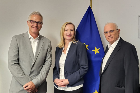 Trzy osoby stoją przodem i patrzą na wprost. Mężczyzna po lewej stronie ma na sobie szary garnitur i białą koszulę, kobieta w środku ubrana jest w ciemny żakiet i białą koszulę, a mężczyzna po prawej stronie ma na sobie ciemny garnitur i białą koszulę. Flaga Unii Europejskiej znajduje się za nimi po prawej stronie.