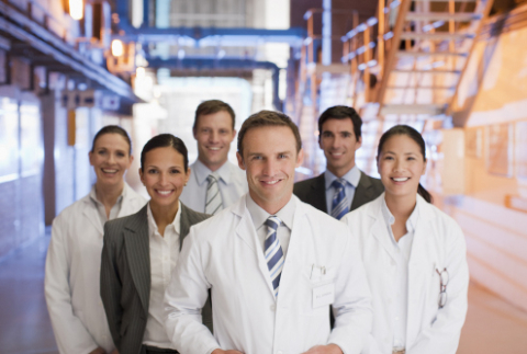 Grupa sześciu osób stojących w przestrzeni przypominającej szpital lub firmę medyczną. W grupie znajdują się zarówno mężczyźni, jak i kobiety. rzy osoby ubrane są w białe fartuchy laboratoryjne, a pozostałe trzy w eleganckie garnitury, co wskazuje na obecność zarówno personelu technicznego, jak i przedstawicieli administracji lub kadry zarządzającej. Wszyscy uśmiechają się do zdjęcia.