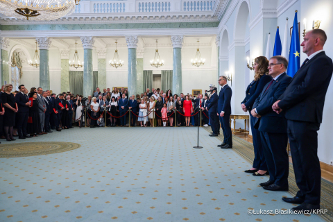 Duża sala z wysokim sufitem i żyrandolami. Na środku mężczyzna przy mikrofonie, zwrócony w stronę grupy po lewej. Po prawej stronie stoi pięć osób w garniturach. W tle tłum ludzi za barierkami. Na ścianie po prawej wiszą flagi Polski i Unii Europejskiej.