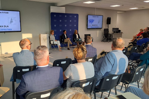 Sala konferencyjna z uczestnikami siedzącymi przodem do sceny. Na scenie trzy osoby i mównica z logo instytucji. W tle dwa ekrany z napisem „Nauka dla społeczeństwa”. Jasne, nowoczesne wnętrze, uczestnicy ubrani elegancko.