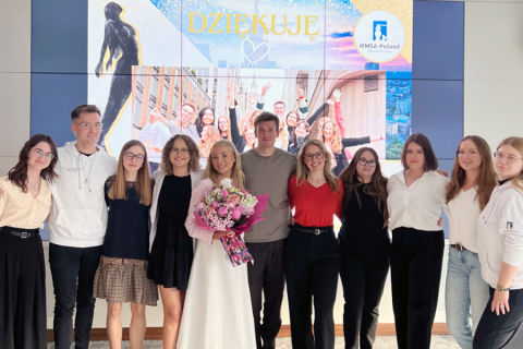 Grupa 11 osób stoi w rzędzie, pozując do zdjęcia. W centrum kobieta w białej sukni trzyma bukiet kwiatów. W tle ekran z napisem „DZIĘKUJĘ”, logo IFMSA-Poland i zdjęciem ludzi z uniesionymi rękami na tle budynków.