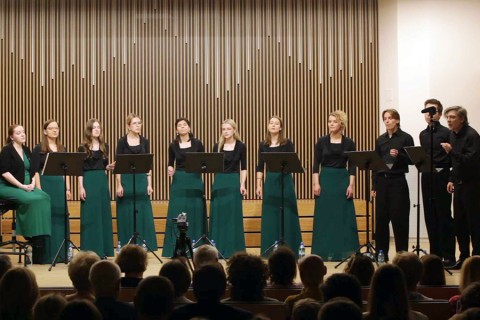 Dziesięć osób stoi na scenie w sali koncertowej, przed pulpitami z nutami. Siedem kobiet w zielonych spódnicach i czarnych bluzkach, trzech mężczyzn ubranych na czarno. W tle drewniana ściana akustyczna z pionowymi elementami. Na pierwszym planie widoczna publiczność siedząca na krzesłach.