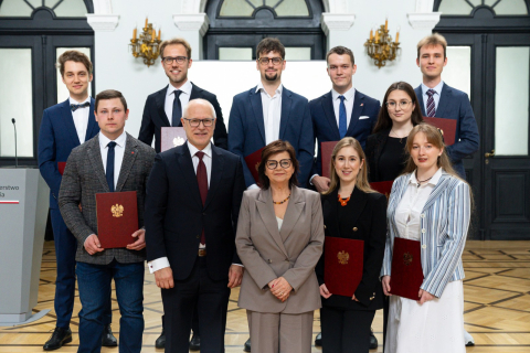 Grupa jedenastu elegancko ubranych osób. Siedmiu mężczyzn, w tym rektor WUM i cztery kobiety, w tym Minister Zdrowia, stoi w dwóch rzędach w sali w budynku Ministerstwa Zdrowia. Większość trzyma czerwone teczki z godłem Polski. Wszyscy uśmiechają się do zdjęcia. W tle widoczne są drewniana podłoga i ciemne drzwi.
