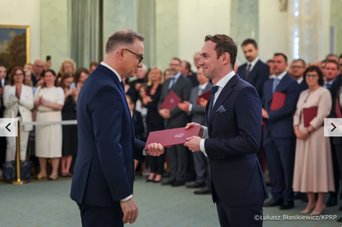 Dwóch mężczyzn w garniturach: prezydent RP oraz naukowiec, wymienia dokument w eleganckim wnętrzu, w tle grupa osób z czerwonymi teczkami.