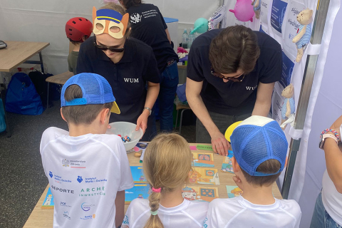 A group of children and adults participate in an educational workshop. Two adults in T-shirts with “WUM” written on them lead the activities at a table with colourful boards. Three children in white T-shirts and blue baseball caps observe. Other people and workshop materials are visible in the background.