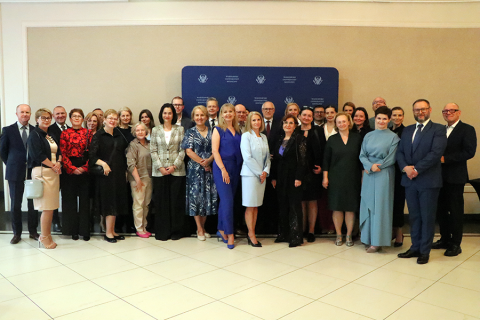 Grupa kilkudziesięciu elegancko ubranych osób pozuje do zdjęcia na tle niebieskiej ścianki z logotypami. Osoby stoją w dwóch rzędach, większość w garniturach i sukienkach. Tło beżowe, podłoga z jasnych płytek.