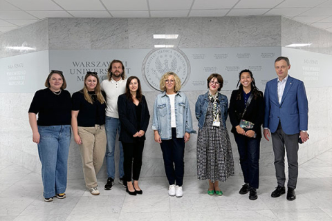 Eight people stand in a row against a light-coloured marble wall with the inscription “Warsaw Medical University”. Other inscriptions and the university's logo are also visible. The participants are wearing outfits of varying degrees of formality - from casual to elegant. They smile for the photograph.