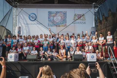 Grupa uczestników wydarzenia sportowego stojąca na scenie ubrana w koszulki z numerami startowymi. Część osób trzyma dyplomy lub medale, inni machają do zdjęcia. 