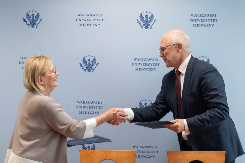 Kobieta, dyrektor NIO i mężczyzna, rektor WUM stoją naprzeciwko siebie i podają sobie ręce. Jedna ubrana w beżowy sweter, druga w ciemny garnitur z białą koszulą i bordowym krawatem. W tle niebieska ściana z logo i napisem „Warszawski Uniwersytet Medyczny”. W rękach mają dokumenty. Kobieta jest ubrana w jasnym sweter, mężczyzna w ciemny garnitur.