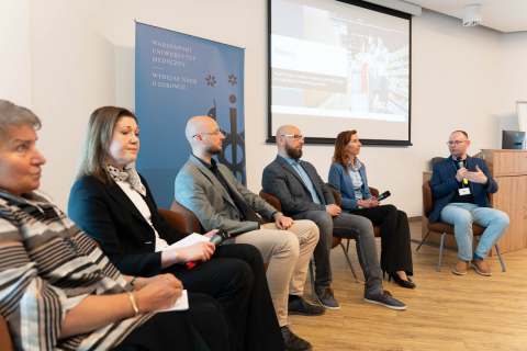 Six people sit in a row on chairs during a panel discussion. Five of them are holding microphones. In the background is a blue banner with the words “Warsaw Medical University, Faculty of Health Sciences” and a screen with the presentation “Panel 1.” The room has light-colored walls and a wooden floor.