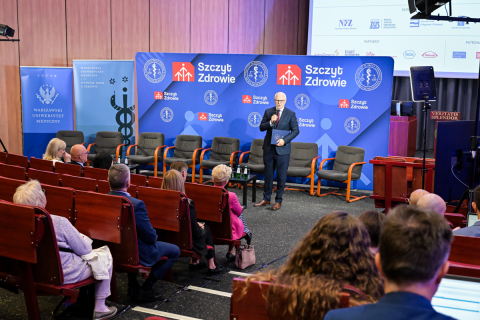 Konferencja „Szczyt Zdrowie 2025” z 12 czerwca 2025 roku. Na pierwszym planie uczestnicy siedzący na krzesłach, w tle scena z prelegentem i niebieskimi banerami z logotypami sponsorów. Na ekranie za prelegentem widoczne są informacje o wydarzeniu, w tym data, tytuł i logotypy partnerów.