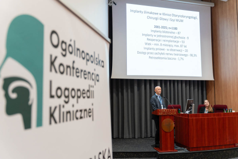 Sala konferencyjna z banerem „Ogólnopolska Konferencja Logopedii Klinicznej”. Na scenie mężczyzna przy mównicy „VERITATIS SPLENDOR”. W tle ekran z prezentacją o implantach ślimakowych w Klinice Otorynolaryngologii WUM.