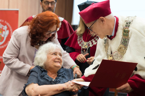 Starsza kobieta na wózku inwalidzkim odbiera dyplom od mężczyzny w czerwonym birecie i białym płaszczu akademickim. Obok niej stoi osoba w jasnej marynarce. W tle widoczne są inne osoby w togach oraz fragment czerwonego baneru z herbem uczelni.