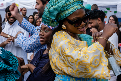 Grupa osób tańczących w trakcie festiwalu. W centrum kobieta w żółtej sukni z wzorem i turkusowym nakryciu głowy, z biżuterią. Na oczach ma ciemne okulary, w uszach złote kolczyki. W tle kolorowo ubrane osoby, namioty i dekoracje festiwalowe.