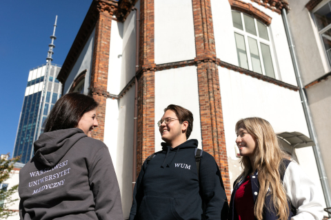 Trzy osoby stoją przed budynkiem. Osoba z lewej ma szarą bluzę z napisem „Warszawski Uniwersytet medyczny". W tle widać budynek. 