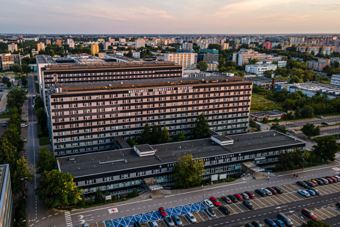 Zdjęcie szpitala przy ul. Banacha z lotu ptaka. Na dachu dużego, jasnego budynku widoczny napis „Warszawski Uniwersytet Medyczny”. Przed nim parking z autami i niebieskimi miejscami dla osób z niepełnosprawnościami. W tle panorama Warszawy z blokami i zielenią.