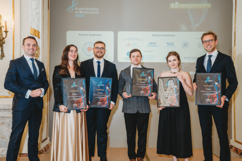 Sześć elegancko ubranych osób. Wszyscy uśmiechają się do zdjęcia. Pięć z nich trzyma w rękach dyplomy. Stoją w jasnym pomieszczeniu z wysokimi sufitami, na ścianach wiszą ozdobne świeczniki. Za osobami widać ekran multimedialny.